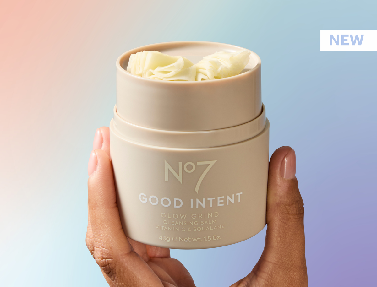 New No7 Good intent Glow Grind Cleansing Balm. Product packaging shown in the hand of a model. Pastel colour ombre background featuring pink, purple and blue shades.