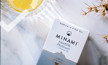 Серо-белая упаковка Омега-3 с витамином Д лежит на мраморном столе рядом со стаканом воды с лимоном.