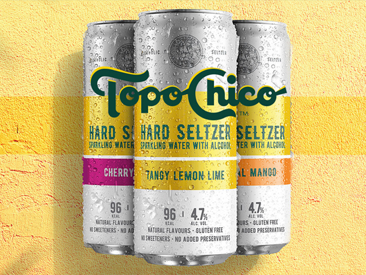 Three Topo chico cans set ahainst a yellow painted wall, lemon and lime flavour, mango flavour and cherry flavour