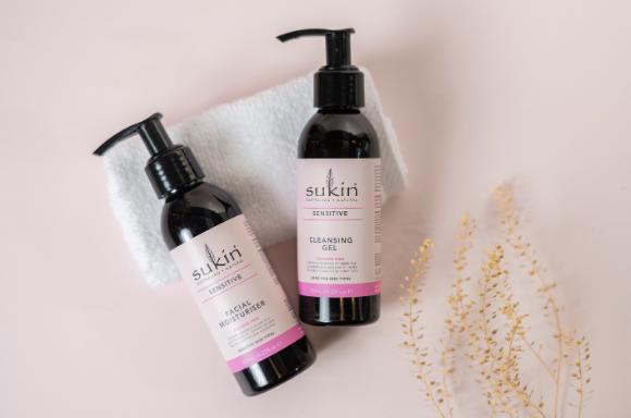 Sukin sensitive facial moisturiser and cleansing gel on a towel and pinky beige background with a few stems of a plant for decoration.