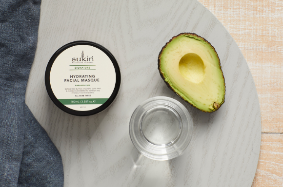 Sukin hydrating facial masque with half and avocado and glass of water for decoration.
