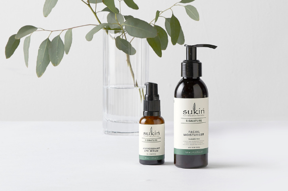 Sukin Siganture facial moisturiser and antioxidant eye serum on background with jug of water behind it and a plant leaf.