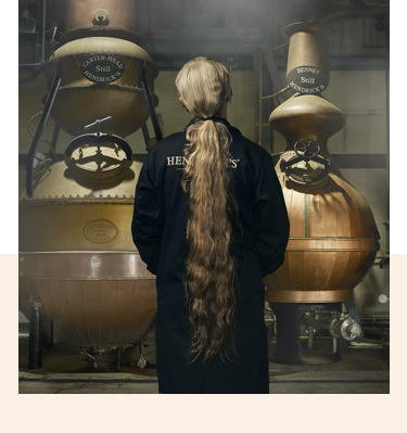 A man with long hair, wearing a black robe, stood looking at two whisky making machines