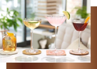 Four cocktails of various colours in a line, each contain a piece of fruit, a window is present in the background which is providing warm lighting