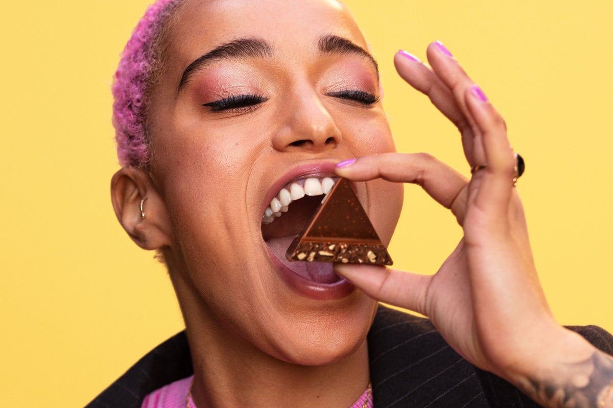 Woman with pink hair eating one piece of a Toblerone in front of a Yellow background