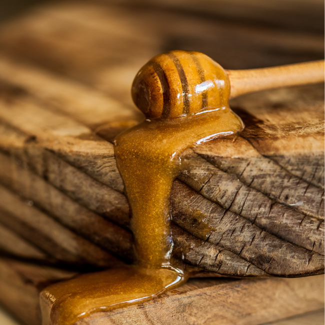 A wooden honey dipper laying on a wooden table and manuka honey is slowly flowing down the table.