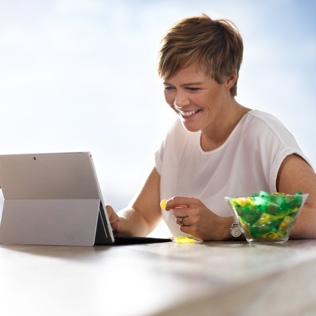 A happy woman working on the laptop and eating sweets. A bowl full of sweets stands next to her.
