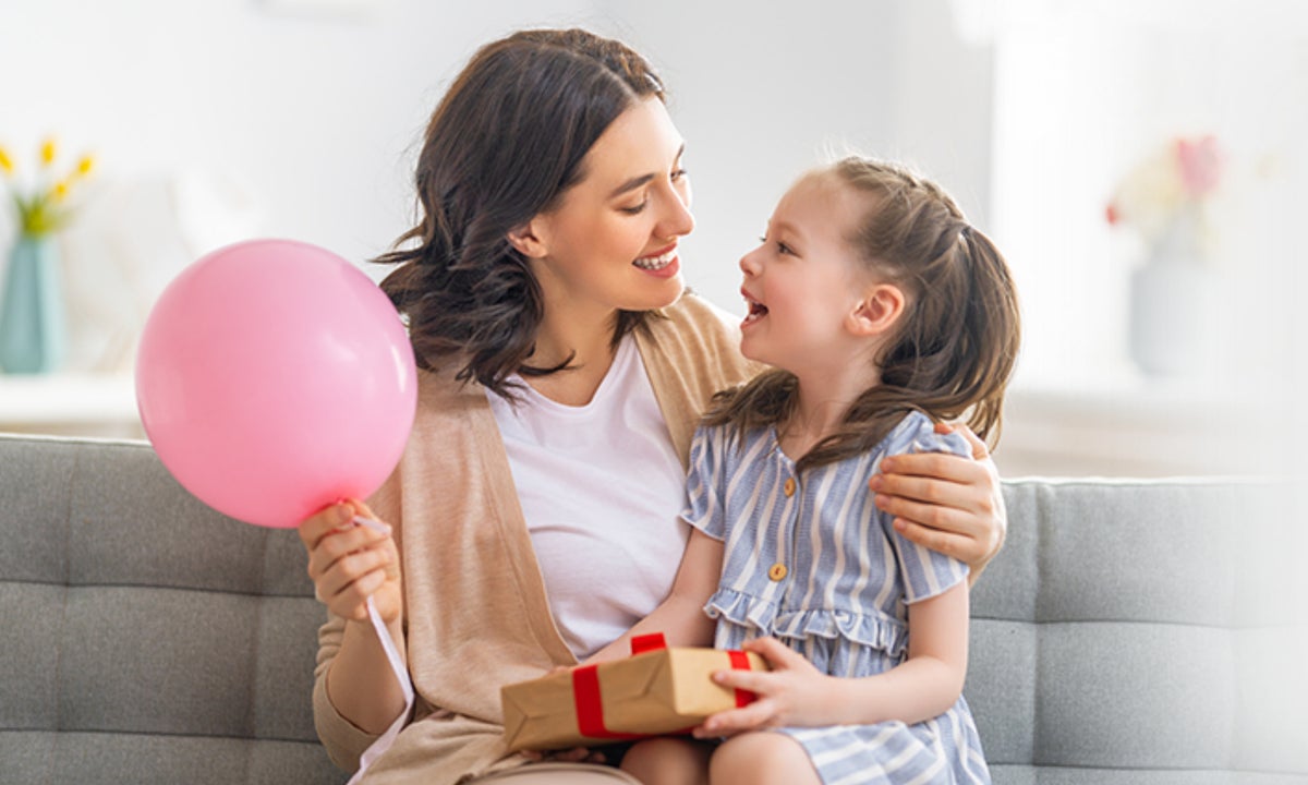 Mama i jej córka uśmiechają się, mama trzyma różowy balon a dziewczynka trzyma w rękach prezent.
