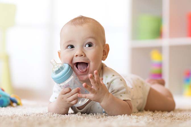 Uśmiechnięte dziecko leży na dywanie, trzymając butelkę z mlekiem Nan Nestle.