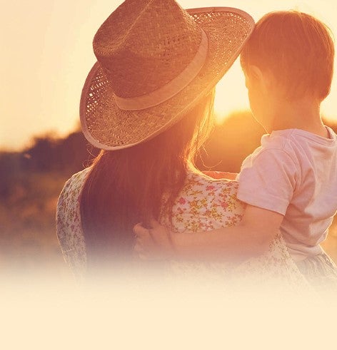 Over ons - Een vrouw die haar peuter vasthoudt. Ze kijken naar de zonsondergang in de natuur.