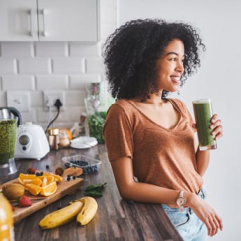 Una giovane donna in piedi, in cucina, che beve uno smoothie verde. Il tavolo dietro di lei è coperto da frutta e verdura.