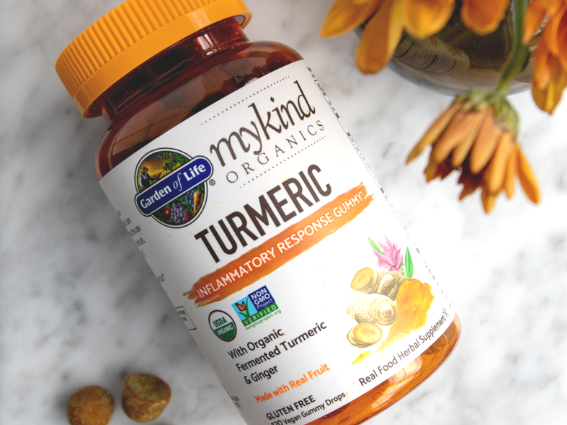 Una confezione di curcuma mykind di Garden of Life si trova su un tavolo bianco ed è circondata da fiori arancioni.