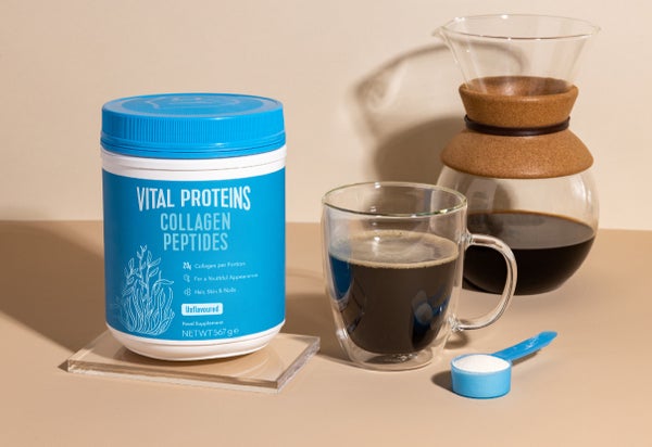 Vital Proteins Collagen Peptides and a blue scoop filled with collagen powder standing on the table next to a cup of coffee.