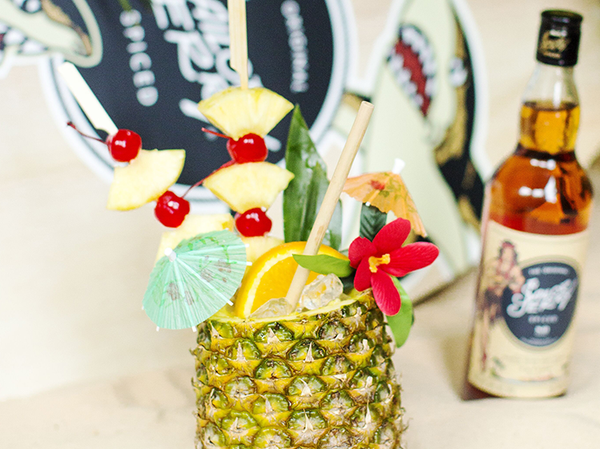 A cocktail in a carved pineapple, it is filled with various fruits, on the right-hand side is a bottle of Sailor Jerry spirit