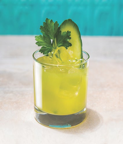 A yellow cocktail in a glass with ice and, cucumber and leaves also present