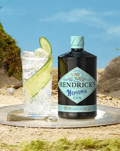 A bottle of Neptunia gin is placed beside a glass containing the gin, it also contains ice and a long slice of cucumber.