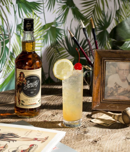 A bottle of Sailor Jerry whiskey with a cocktail next to it containing ice, lemon and a cherry, with an image of an older gentleman smoking a pipe in the background