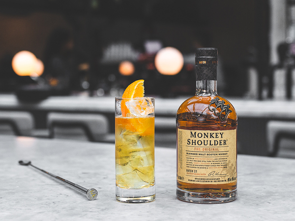 A cocktail containing ice, an orange slice, and a stirrer, placed besides a bottle of monkey shoulder whisky