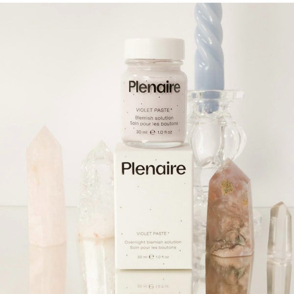 Plenaire Violet Paste blemish solution on the product box on a glass table surrounded by crystals and a candle holder with swirled blue candle against a white background. Visit our instagram