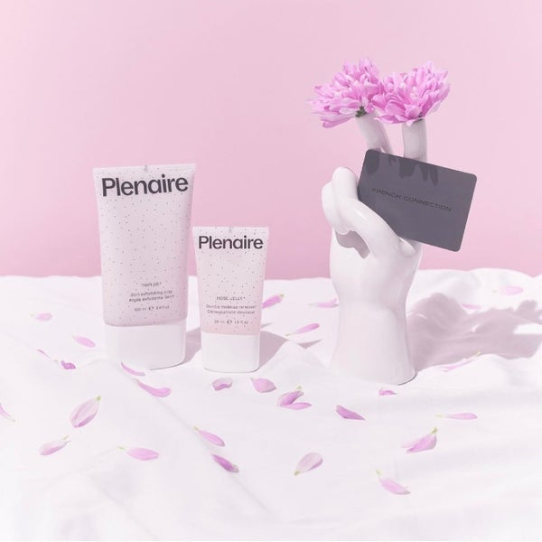 Plenaire Tripler 3in1 exfoliating clay and Plenaire Rose Jelly Gentle Makeup Remover stood on a white sheet with rose petals against a pink backdrop next to a white hand sculpture with two pink flowers on the ends of the two fingers with a Black French Connection card. Visit our instagram
