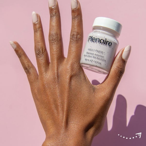 An opened hand with fingers spread apart whilst holding Plenaire's Violet paste blemish solution against a pink background. Visit our instagram