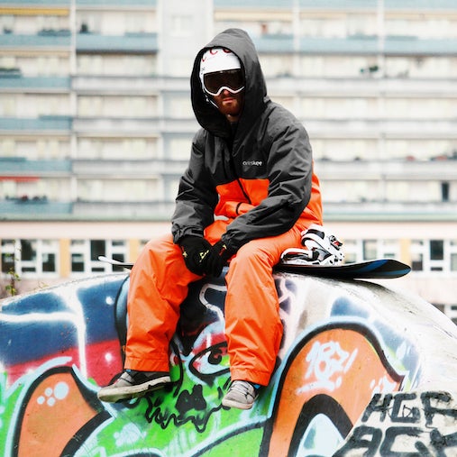 Visit Oneskee Instagram. Man wearing our black and orange snowsuit and white goggles, sat on graffitied wall.