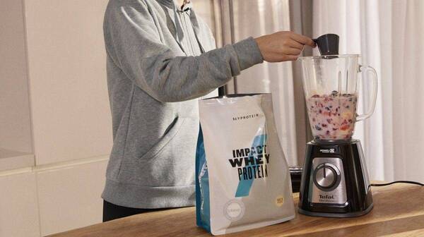 A person adding a scoop of Impact Whey Protein to a blender full of fruit and milk.
