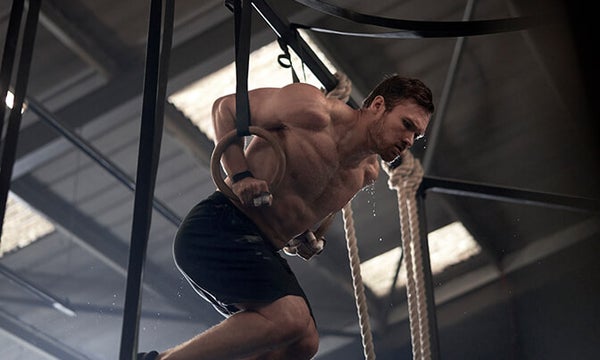 A man working out topless with black gym shorts on, using gymnastic rings with sweat dripping off.
