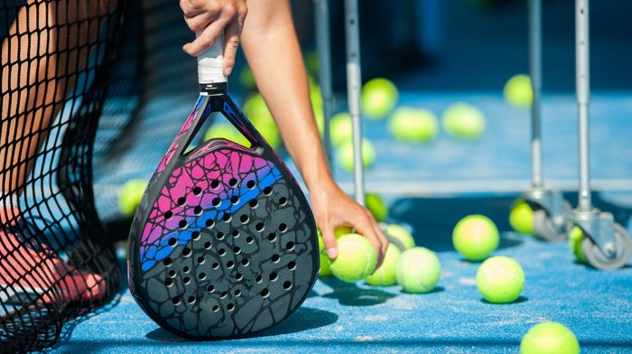 A person holds a padel bat while picking up a ball on an enclosed court.