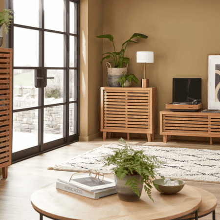 Small round table with a plant on top. In the background, a rug and small cabinets with another plant on.