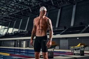 Swimmer Adam Peaty stood in front of a pool wearing Speedos, a swim cap and goggles.