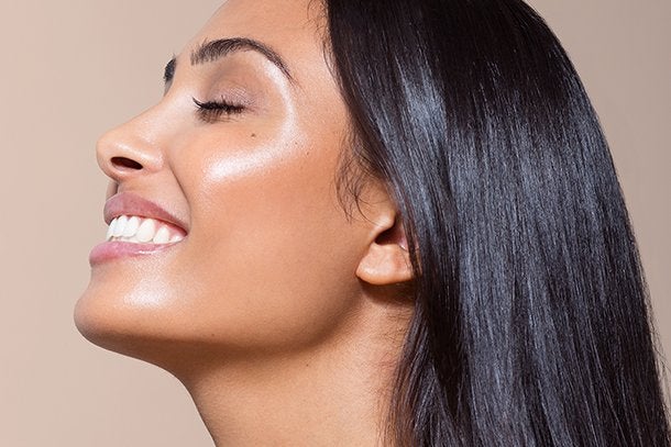 a side profile of a model smiling with glowing skin, pink lipstick and shiny straight black hair on a beige studio background.