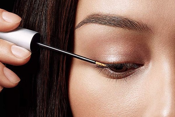 An extreme close up of a light-skinned model with dark features applying an eye lash serum to her top lashes while looking down