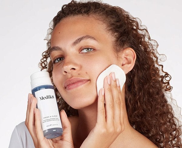 A medium shot image of a female model with curly hair applying Medik8's Pres and Glow tonic on her face with a cotton pad.
