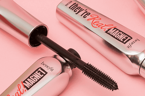Two of Benefit’a They're Real Mascaras, one opened and one closed, in a studio setting on a pale pink background at an angle.