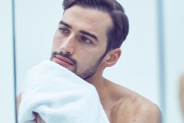 A medium shoot of male model with dark hair wiping his face in front of a mirror with a white towel in a bathroom setting.