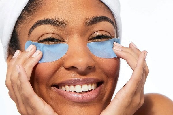 Medium shot of darked skinned model smiling at the camera wearing a head towel and blue eye mask on her undereyes, in a bathroom setting. 