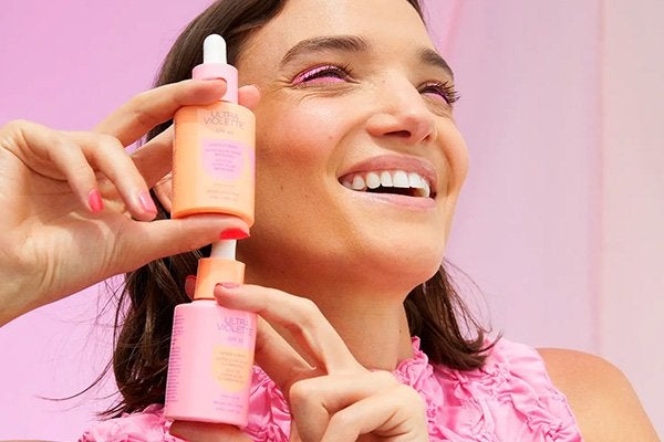 A model wearing a floral pink top, dark hair and pnk eyeliner holding two bottles of the ultra violette glow drops. shot in a studio with a pink background