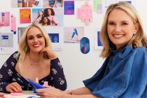 two blond women, one wearing a blue short sleeved denim shirt, sitting at a table in a boardroom with ultra violette branding and imaging in the background