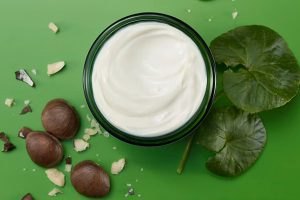 on a green background, there's an open pot of a white cream surrounded by hazelnuts and green leaves