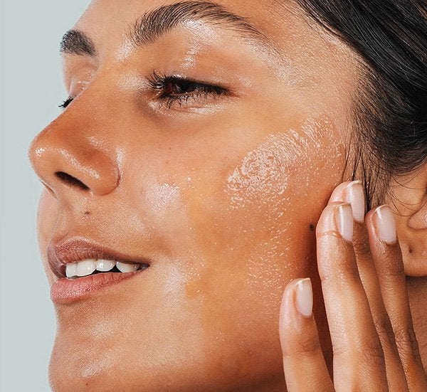 A close up image of a woman rubbing in a barrier-boosting serum into her face as she looks to the side in a studio setting, on a grey background.