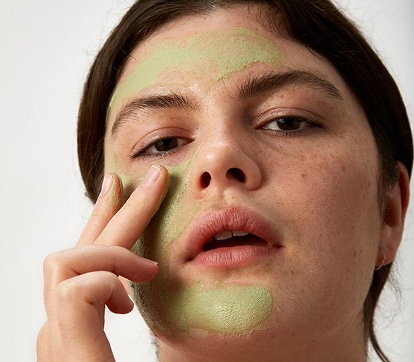 A close up shot of a female model with brunet hair applying a sensitive skin mask on her bare face, in a studio setting.