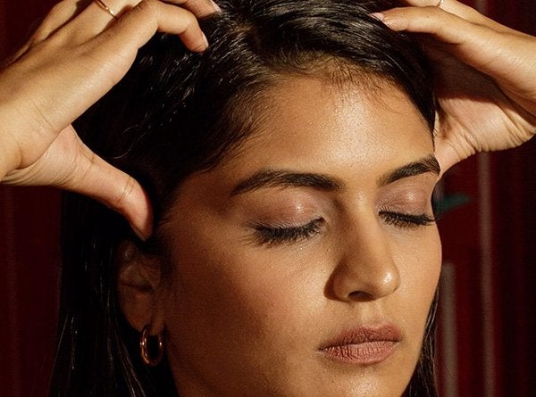 A medium shot of a female model with dark hair massaging her scalp and strands with hair oil as part of her hair oiling ritual.