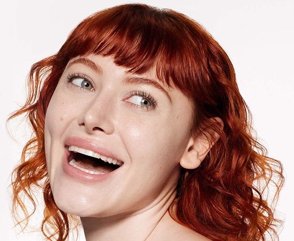 A close up of a model with red hair and arched brows doing a wide grin and looking off to the side against a studio background.
