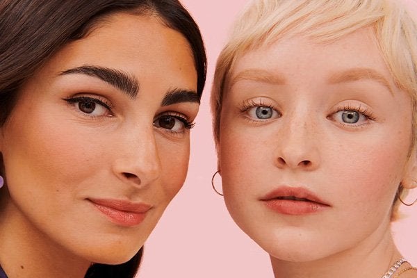 two models one with olive skin dark hair and eyes, and another with pixie blonde hair, fair skin and blue eyes, shot against a baby pink background