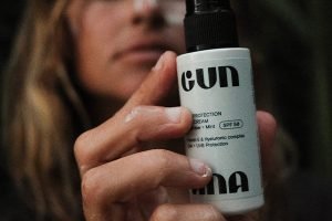 a close up of a gun ana spf mist being held close to the camera. in the background is a blonde haired woman with a swatch of spf on her cheek