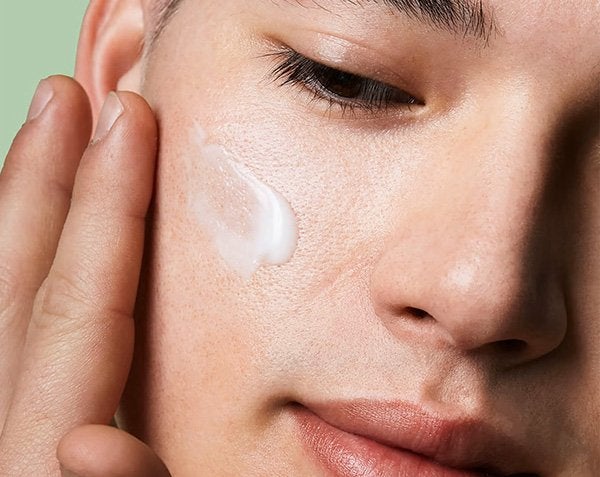 A close up shot of a person applying moisturiser to their right cheek against a pale green background, visible in the top left corner.