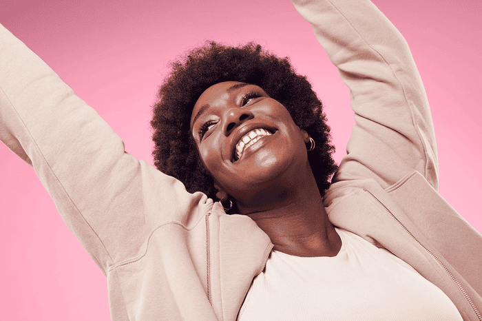 Woman smiling with hands in the air, pink background with afro-textured hair, expressing joy and self-confidence.