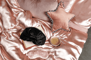 Satin sleep cap on top of pink satin bedding with a pink starred pillow, coffee and book next to it.