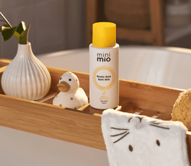 mini mio bath milk on a wooden tray across a bath with rubber duck and a face cloth with a bunny sewed onto it.